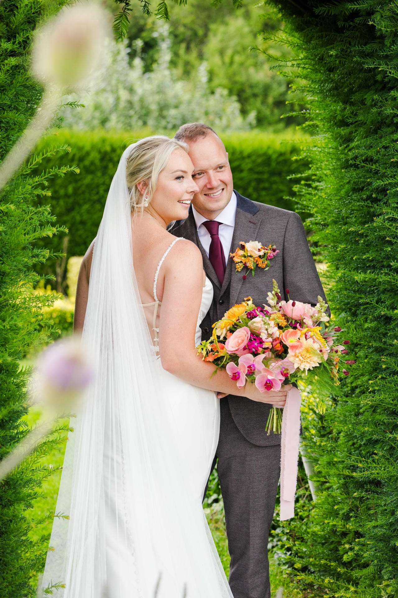 Bride and groom pose in a lush garden, the bride holding a colorful bouquet at The Gardens Wedding venue