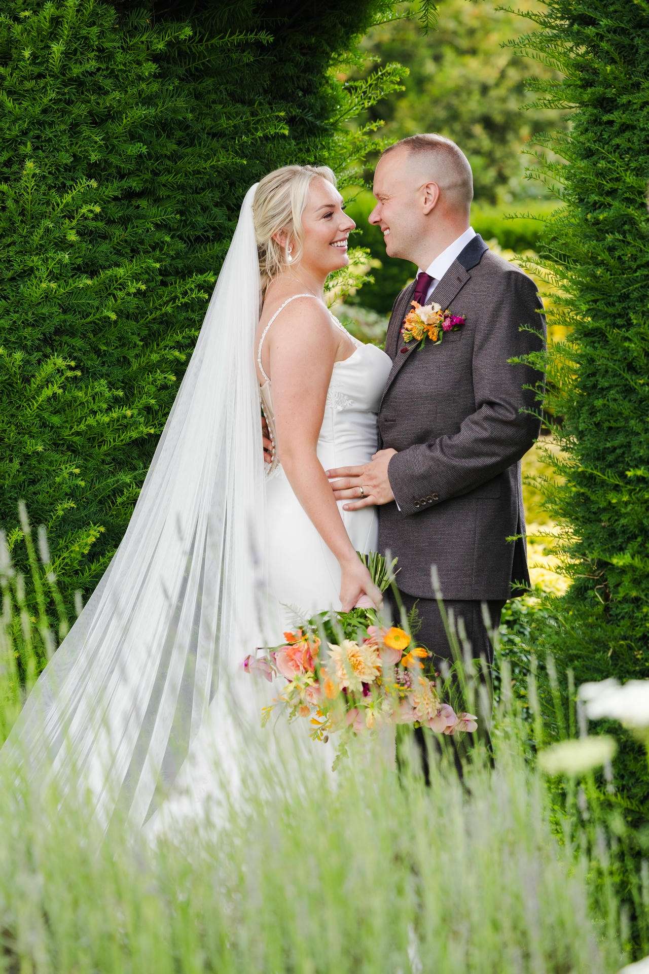 A bride in a white wedding dress and veil stands with a groom in a dark suit, smiling at each other between lush green hedges while the bride holds a colorful bouquet at The Gardens Wedding venue