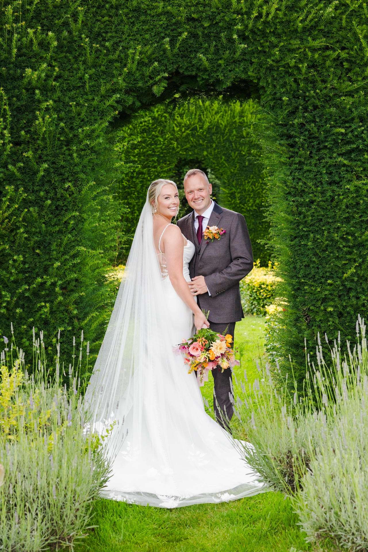 Bride in a white wedding gown with veil and colorful bouquet stands beside a groom in a dark suit under a lush green hedge arch at The Gardens Wedding venue