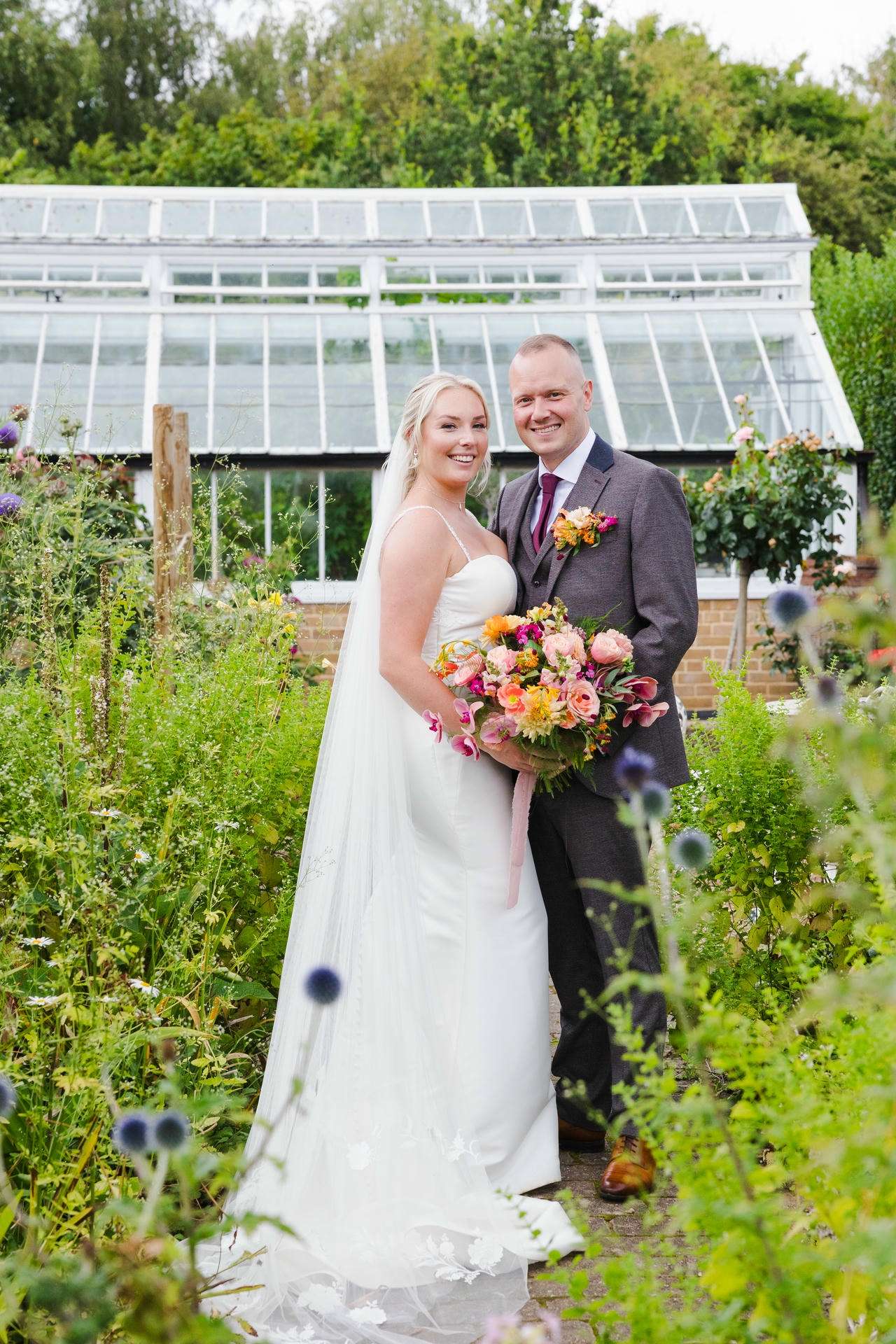 Smiling bride in a white wedding dress with veil and groom in a gray suit pose in a garden, holding a colorful bouquet in front of a greenhouse at The Gardens Wedding venue