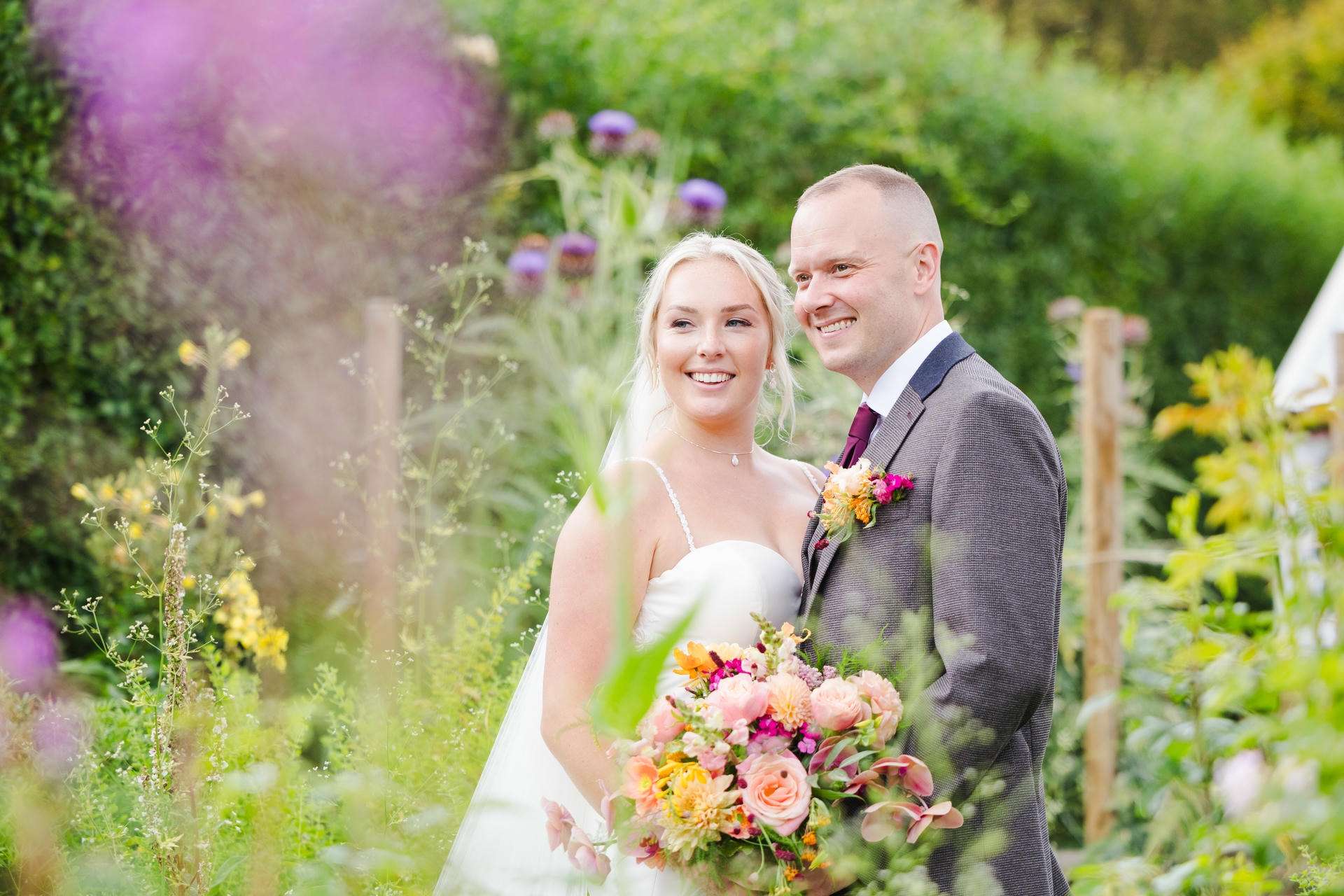 Smiling bride and groom pose in a lush garden, with the bride holding a colorful bouquet at The Gardens Wedding venue