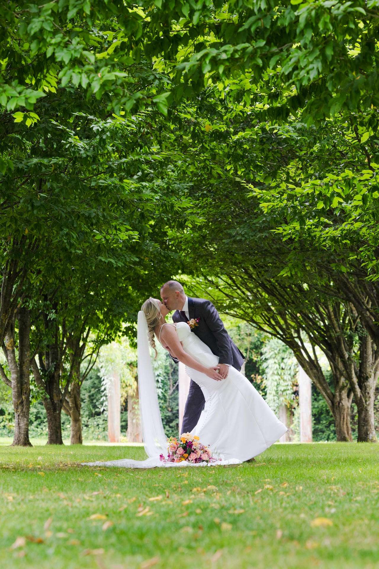 A bride and groom kiss in a tree-lined park, the groom dipping the bride as a colorful bouquet lies on the grass beside them at The Gardens Wedding venue