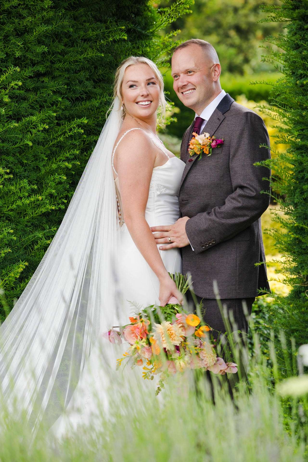 Bride in a white wedding dress and veil stands with a smiling groom in a dark suit outdoors among lush green hedges, holding a colorful bouquet.