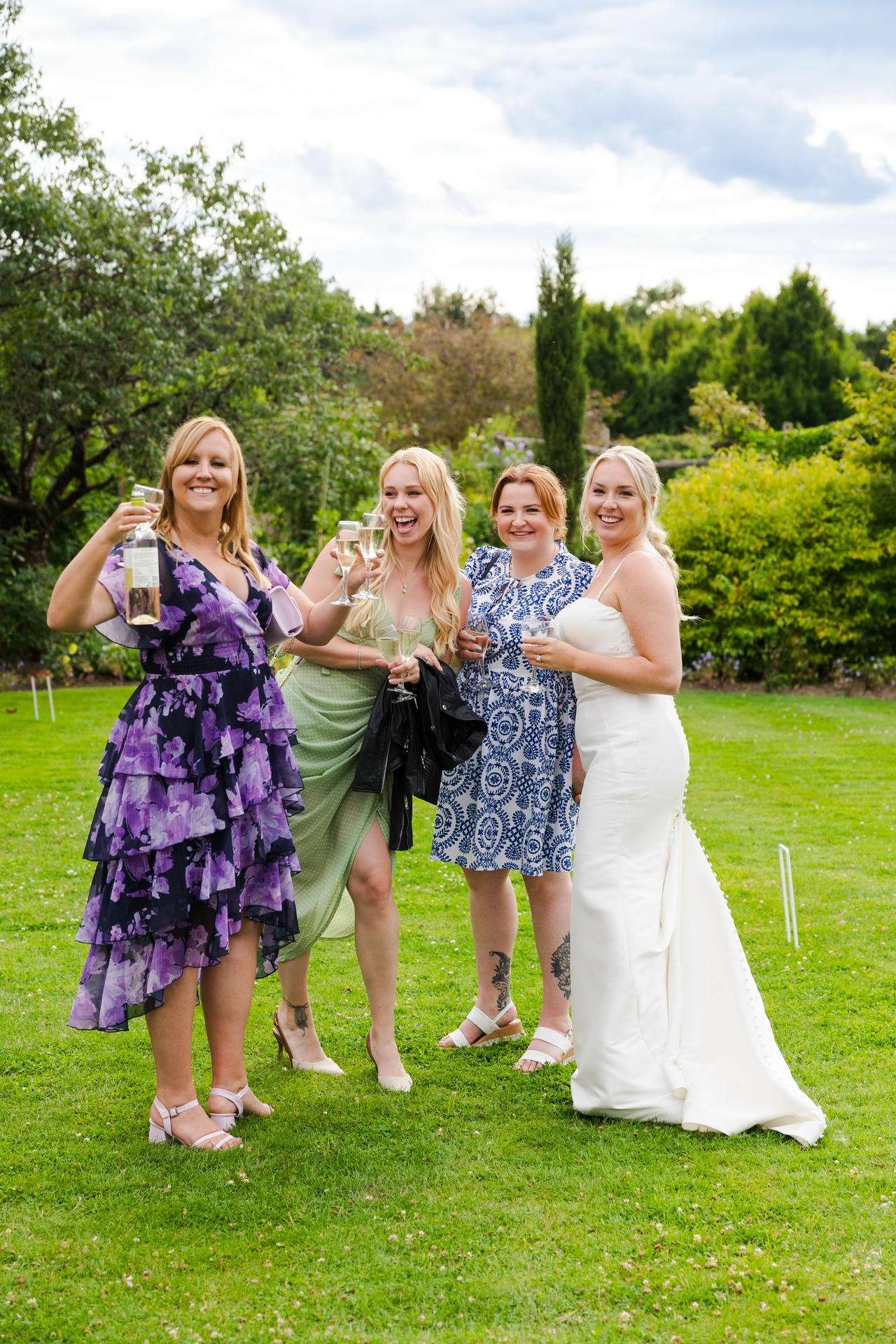Four women in colorful dresses stand on a grassy lawn outdoors, smiling and toasting with drinks, including one in a white wedding dress.