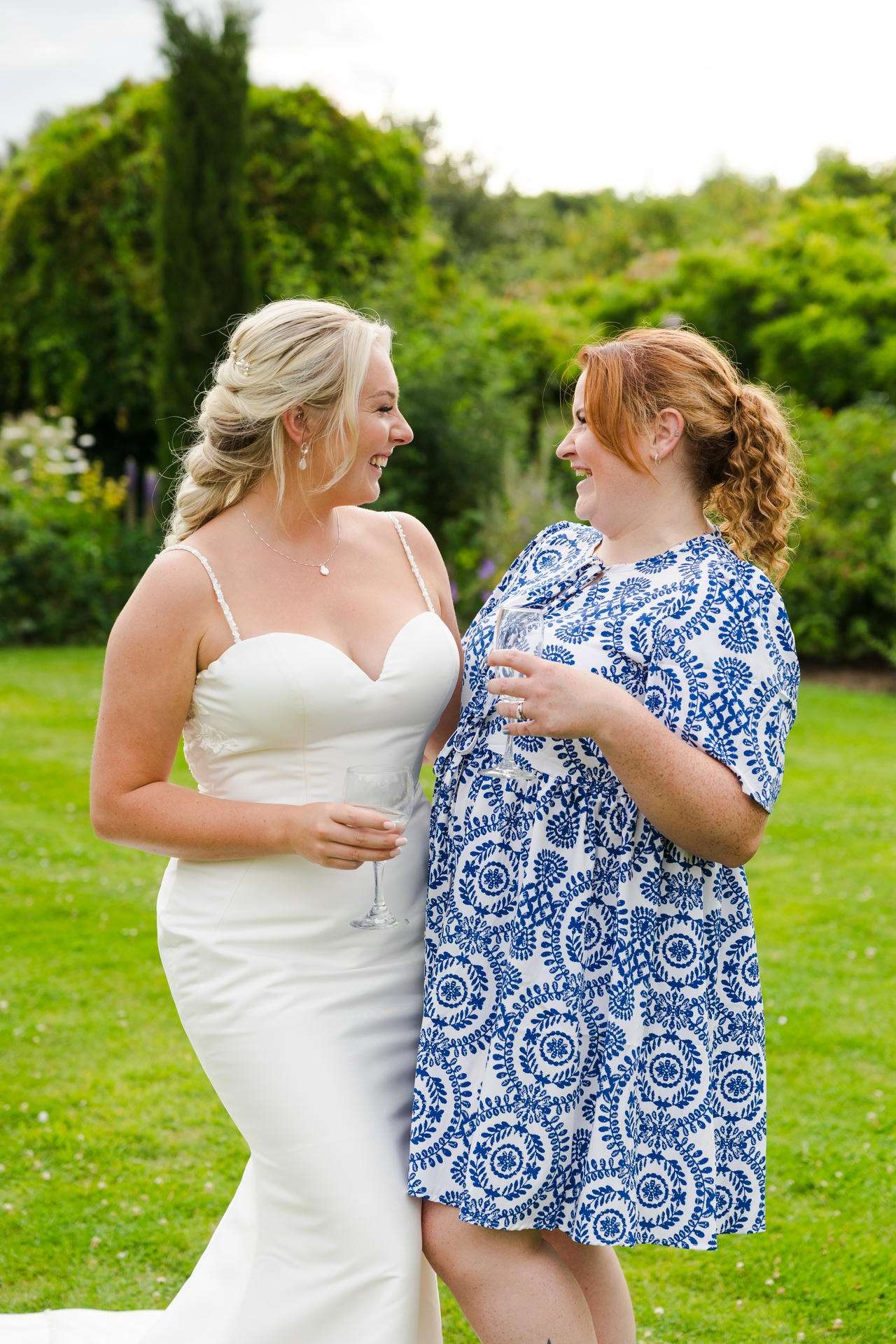 Bride in a white wedding gown and a friend in a blue patterned dress share a smile while holding champagne glasses in a lush garden.