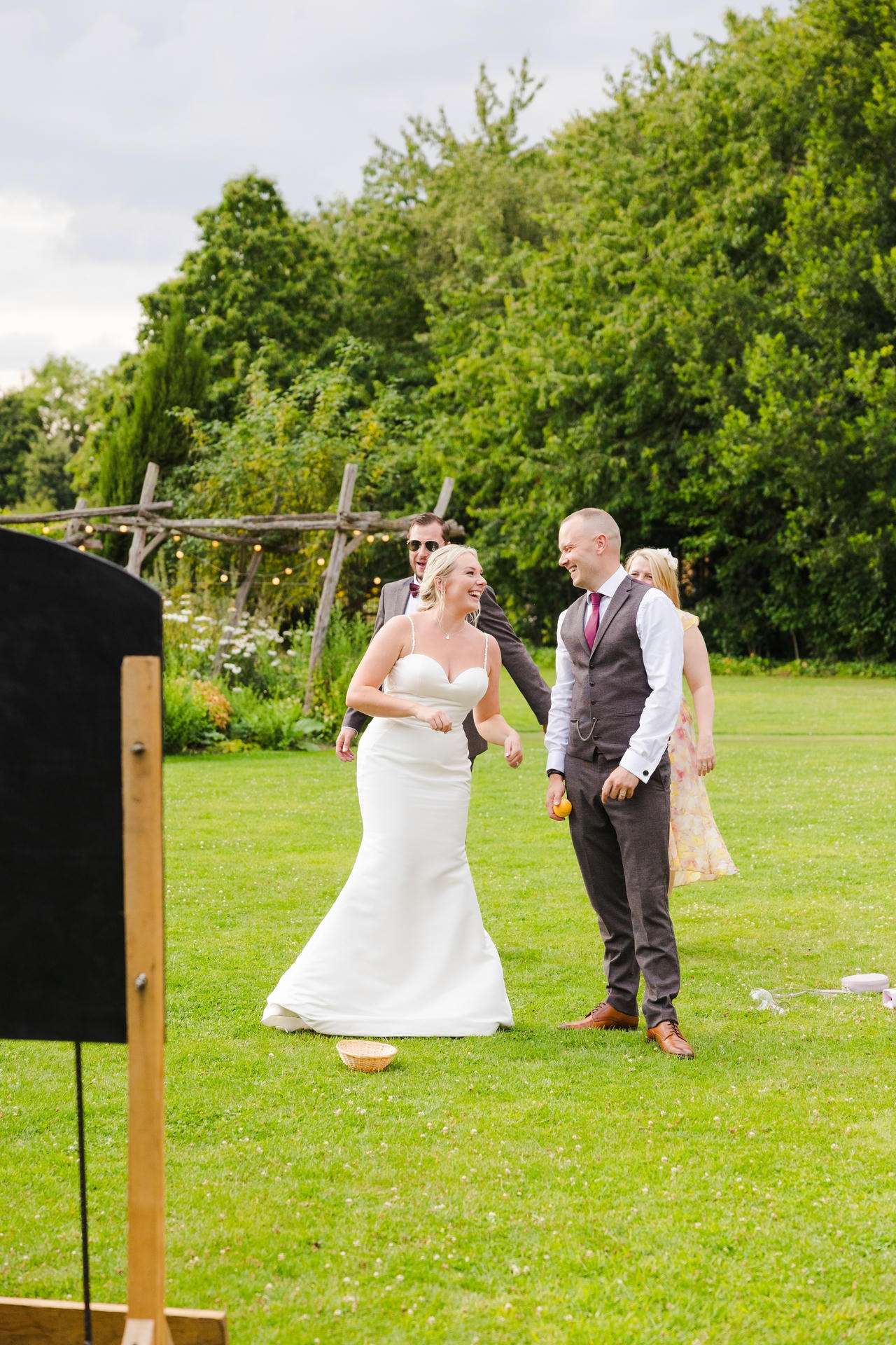 Bride in a white wedding dress and groom in a gray suit share a laugh on a grassy lawn at an outdoor wedding, with guests and lush trees in the background.