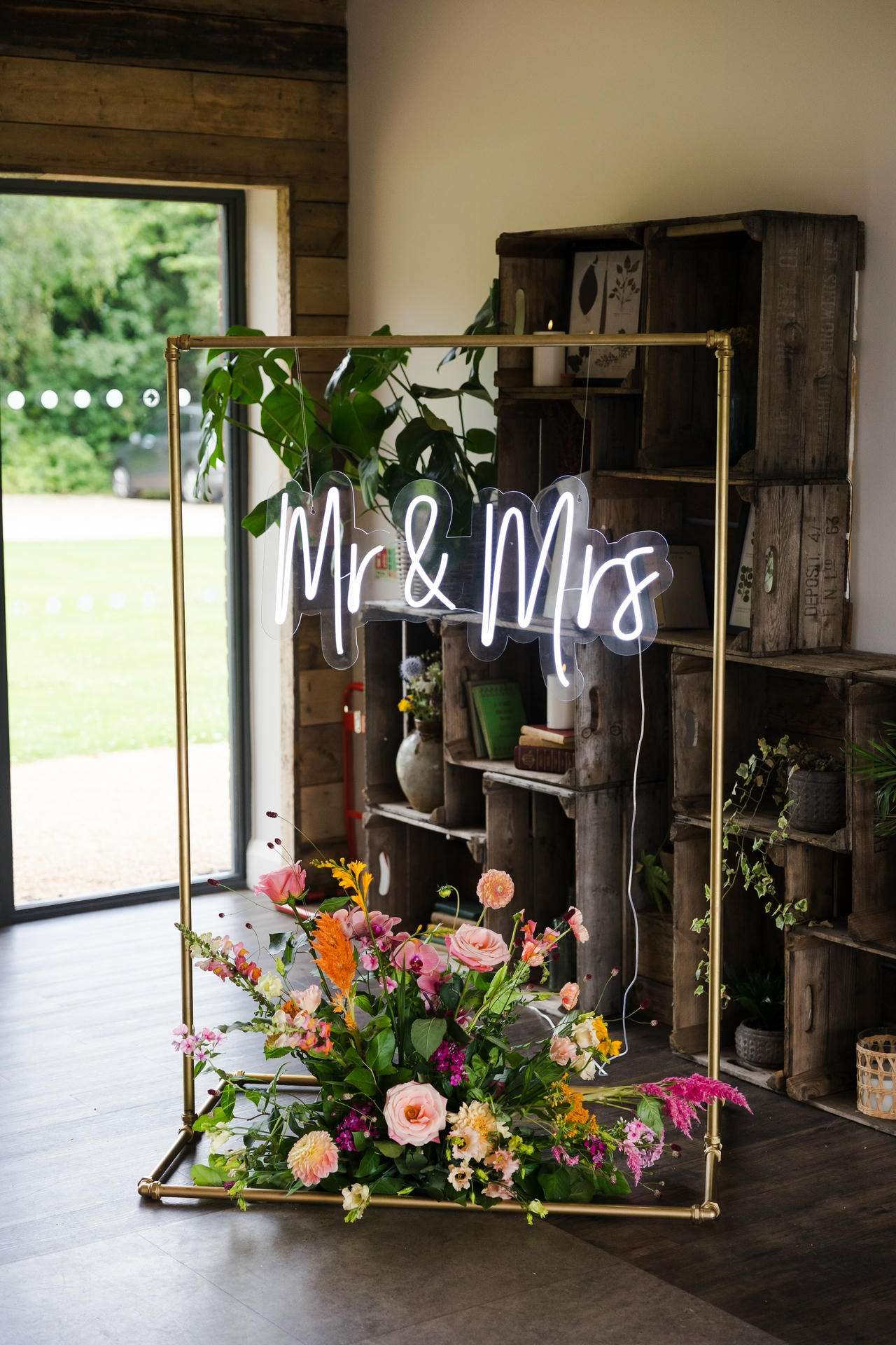 Neon sign reading 'Mr & Mrs' in cursive on a gold frame with a colorful flower arrangement at its base.