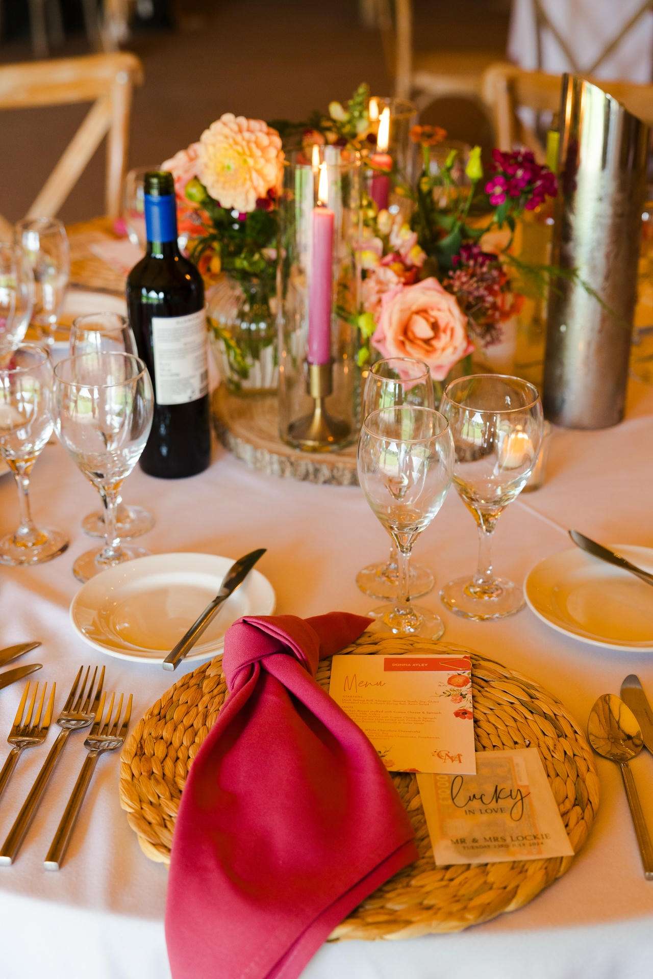 An elegant table setting featuring a floral centerpiece with candles, wine glasses, and a pink napkin on a woven placemat.