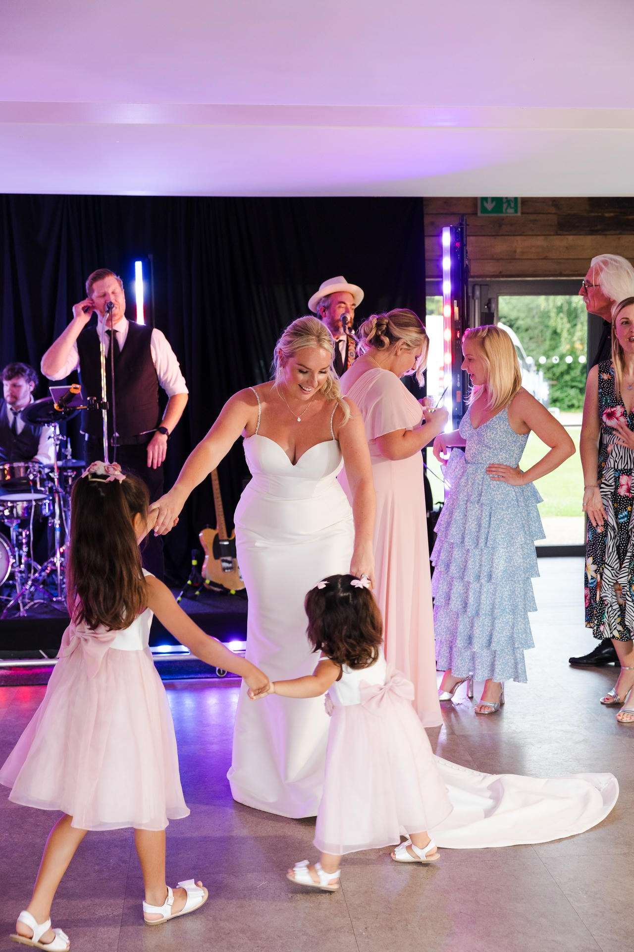 Bride in a white gown dances with two young girls in pink dresses at a wedding reception, with a live band performing on stage in the background.