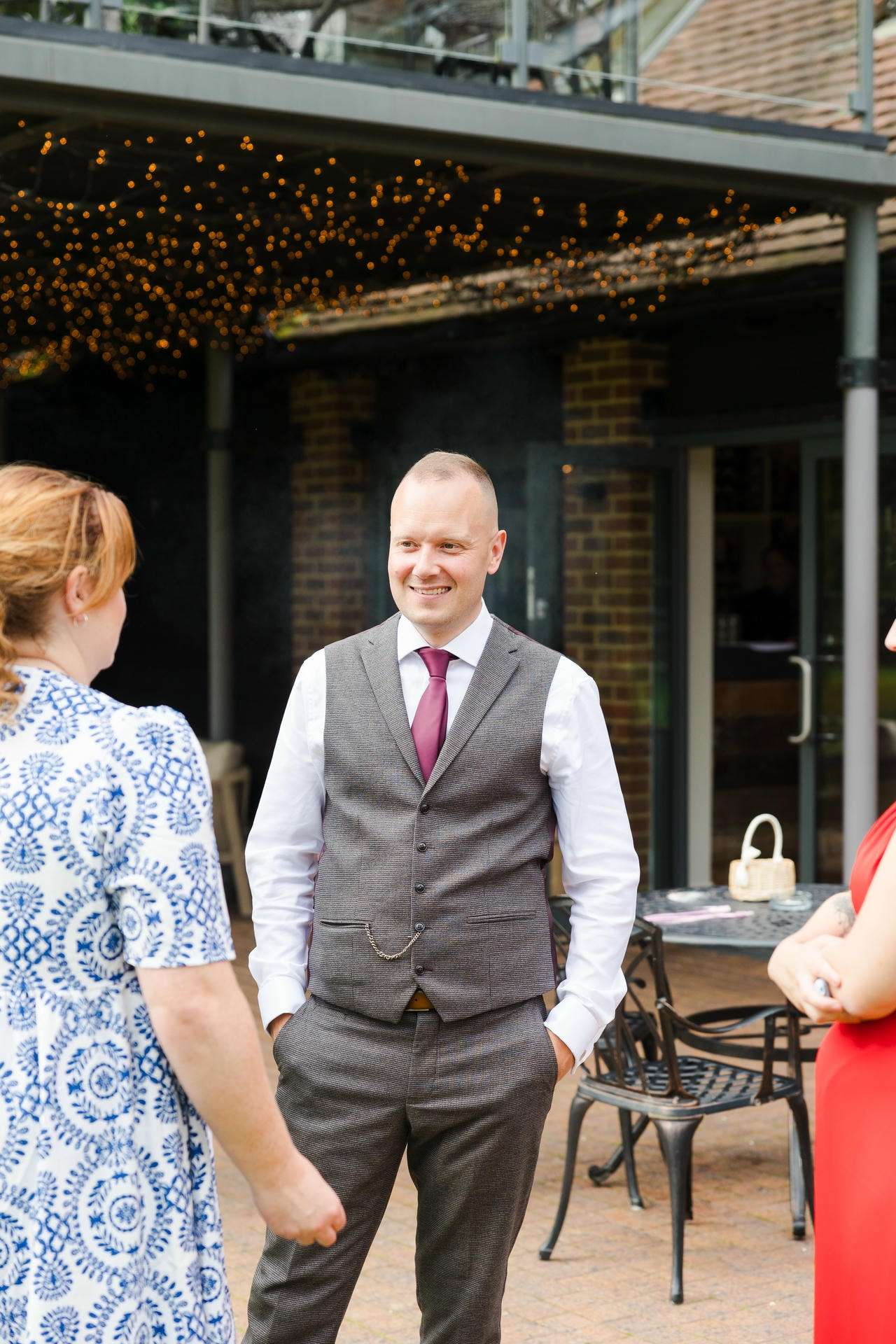 Smiling man in a gray waistcoat and white shirt with a pink tie talks with guests at an outdoor event under string lights.