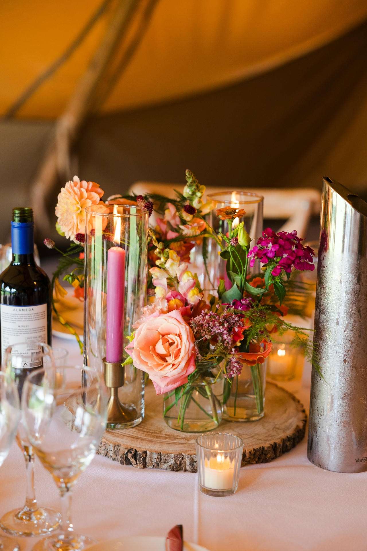 A rustic table centerpiece featuring pink and peach flowers in glass vases, pink candles, a bottle of wine, and glassware on a wooden slab.