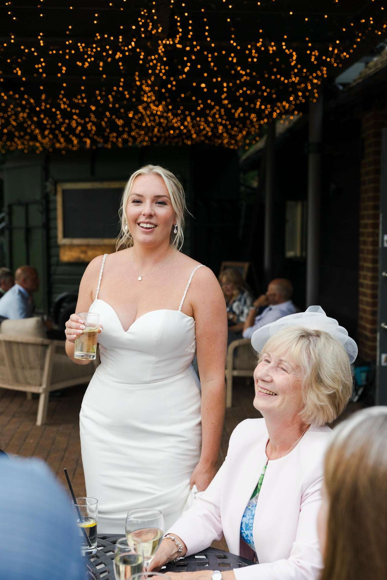 A bride in a white wedding dress holds a glass and smiles, standing beside an older woman in a pale pink outfit at an outdoor reception under string lights.