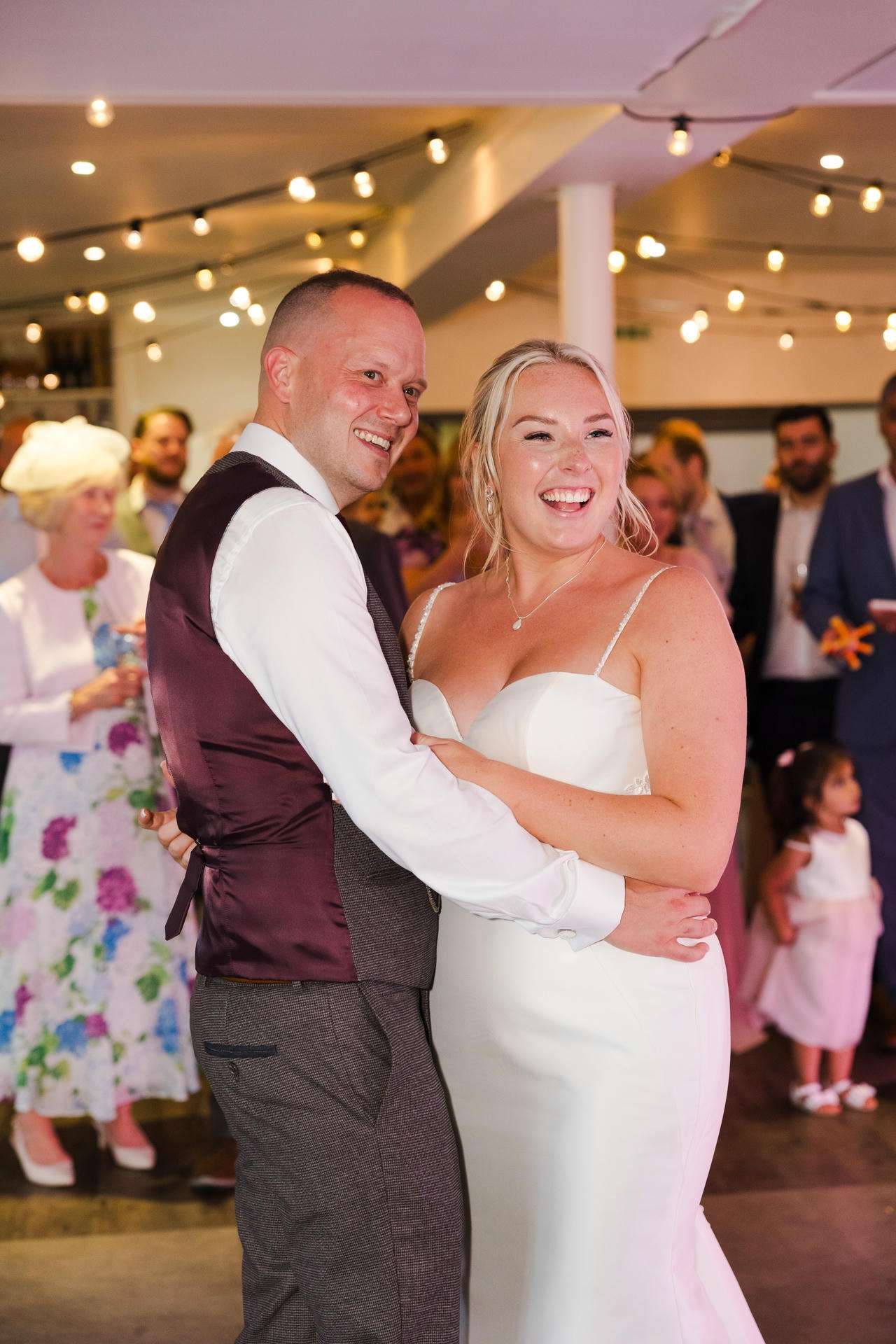 A bride in a white wedding dress and a groom in a maroon vest laugh while dancing together at their wedding reception.