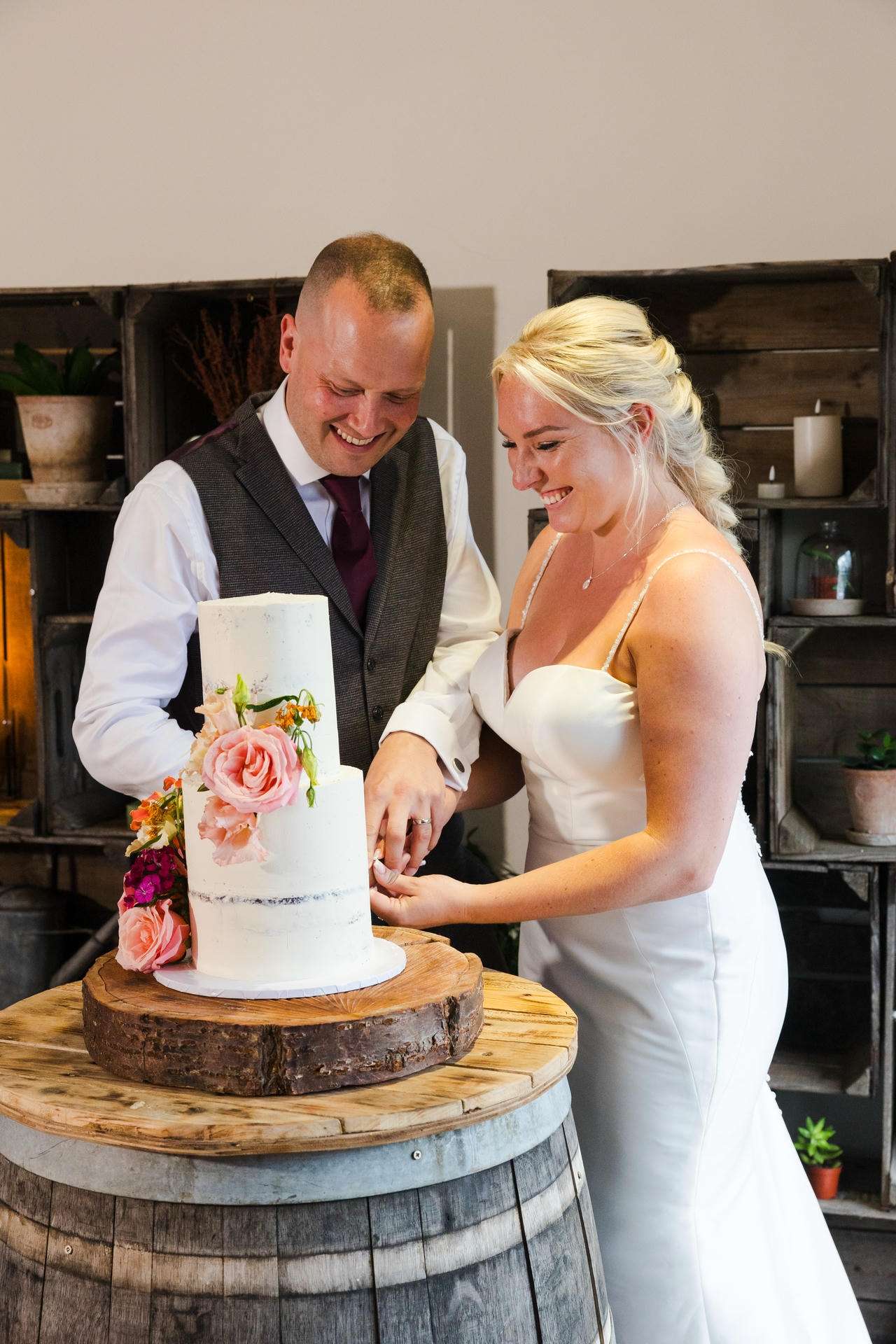 Bride and groom cutting a two-tier white cake decorated with pink and peach flowers on a rustic wooden barrel.