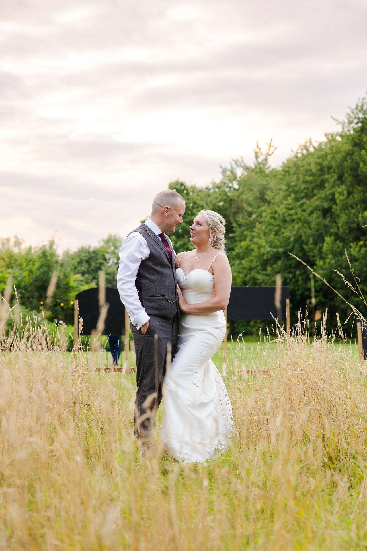 Bride in a white wedding dress and groom in a gray suit stand in a field of tall grass, smiling at each other with trees and a pale sky behind them.