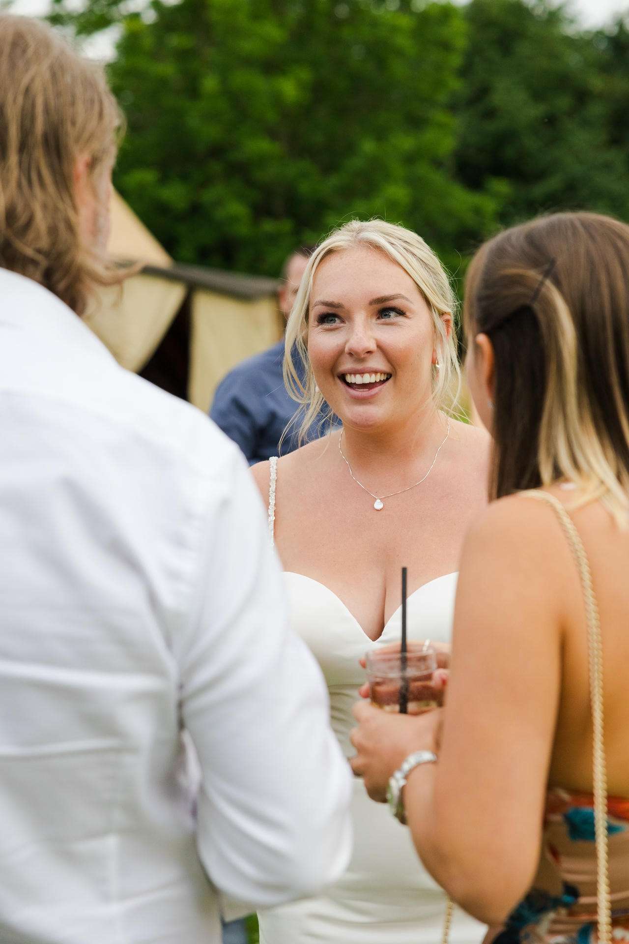 Smiling bride in a white dress chats with two guests at an outdoor wedding.