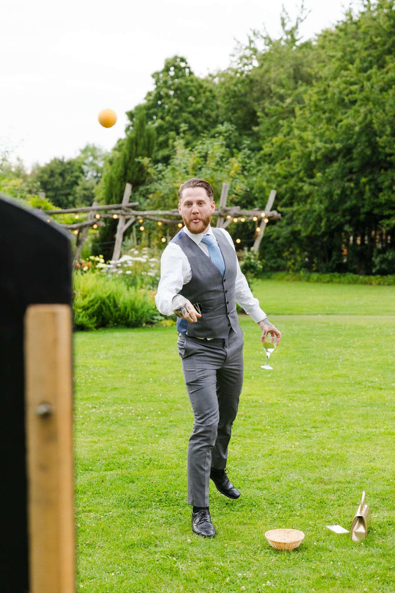 Man in a gray vest and blue tie throws an orange ball across a lush garden, holding a wine glass.