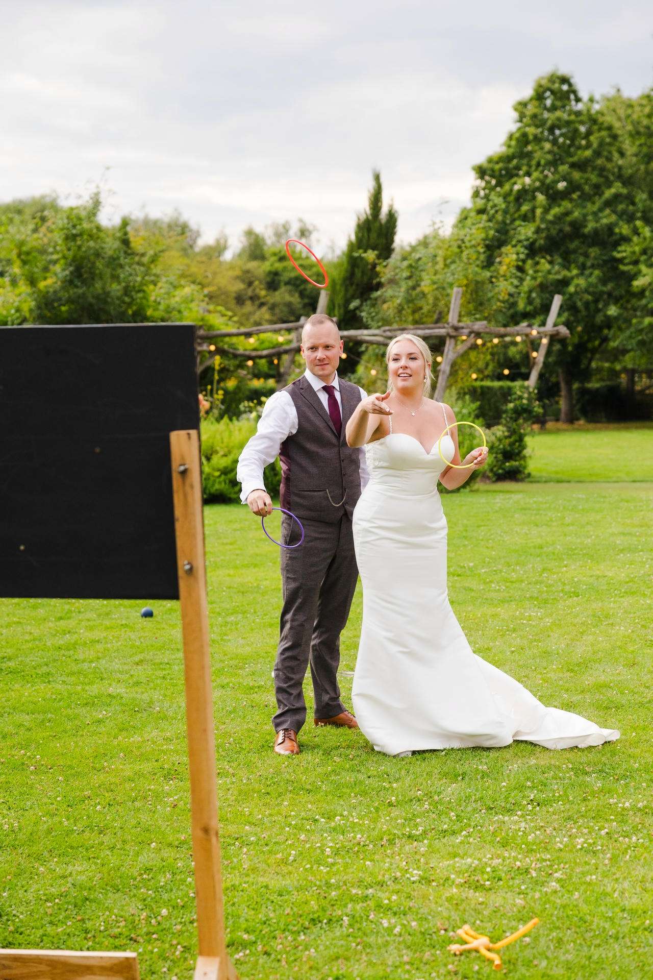Bride in a white wedding dress and groom in a gray vest toss colorful rings on a green lawn in an outdoor garden.