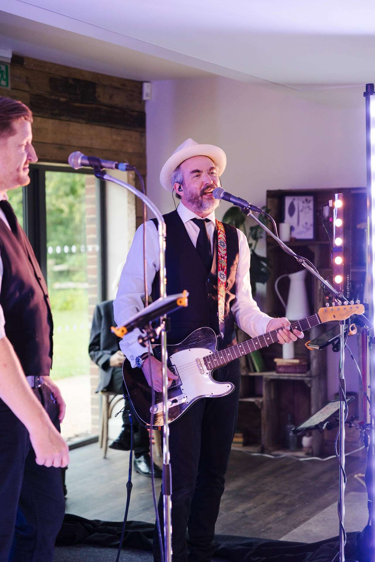 A bearded musician wearing a white hat and vest sings into a microphone while playing a black electric guitar on a small indoor stage.