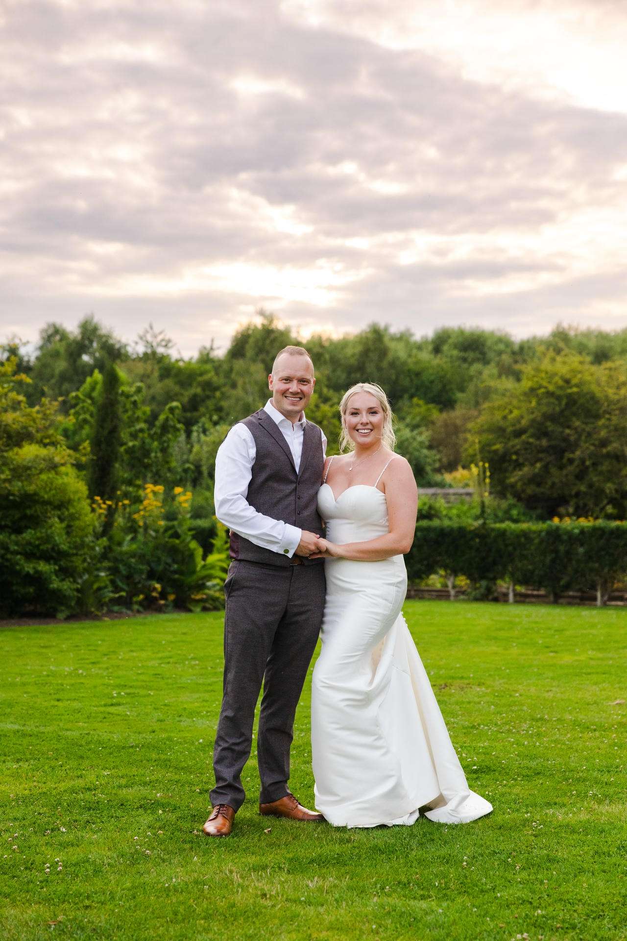 A smiling bride in a white wedding dress and a groom in a gray vest pose together on a grassy lawn with trees behind them.