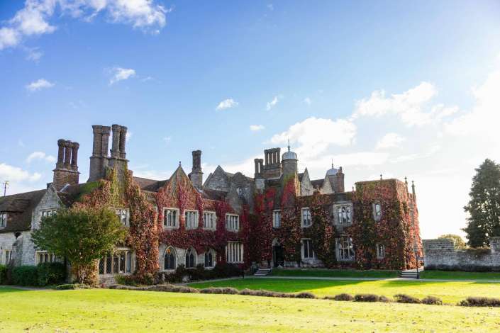 A historic stone building with ivy-covered walls, arched windows, and tall chimneys sits on a green lawn beneath a bright blue sky.