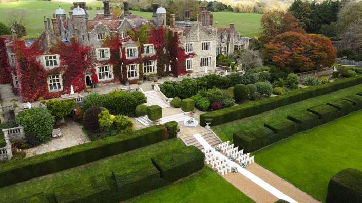 An ivy-covered stone manor with a formal garden and rows of white chairs along an aisle, prepared for a wedding.