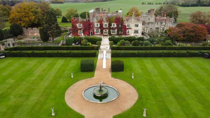 An ivy-covered stone manor with formal, neatly trimmed hedges and green lawns, a central staircase leading to the entrance, and a circular fountain in the foreground.