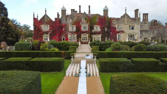 Historic stone estate covered in red ivy with a formal garden and white chairs lined along a central aisle for a wedding.