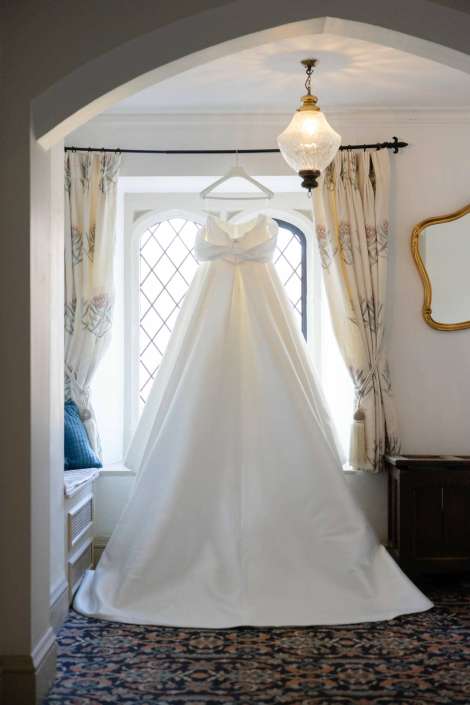 A white wedding dress with a long train hangs on a hanger in a sunlit window alcove, framed by patterned curtains.