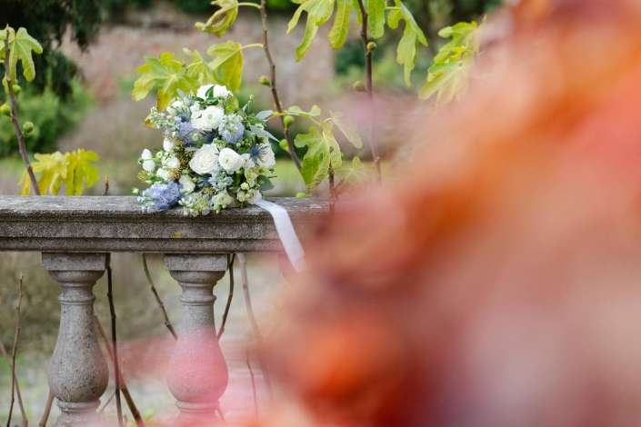 A bouquet of white and pale blue flowers with greenery rests on a weathered stone balustrade in a garden.