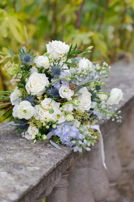 A white and pale blue bouquet with roses, thistles, and greenery rests on a stone ledge outdoors.
