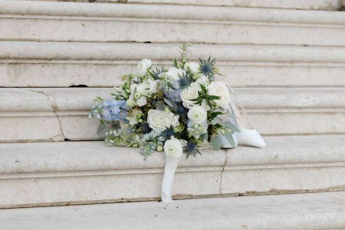 A white bridal bouquet with blue accents rests on pale stone steps beside a white high-heeled shoe.