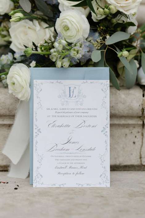 A formal wedding invitation card with decorative borders and script text resting on a light blue envelope, with a white floral bouquet in the background.