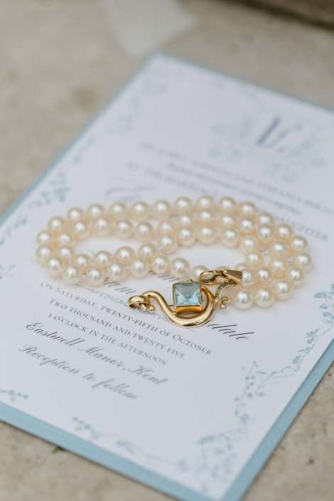 A string of cream-colored pearls with a gold clasp and a blue gemstone rests on an ornate wedding invitation.
