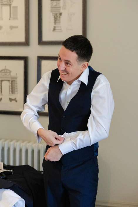A man in a white dress shirt and dark waistcoat smiles as he adjusts his cuff.