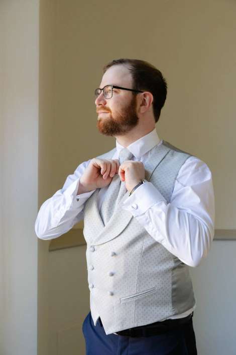Bearded man in a light gray patterned waistcoat and white shirt adjusts his tie.