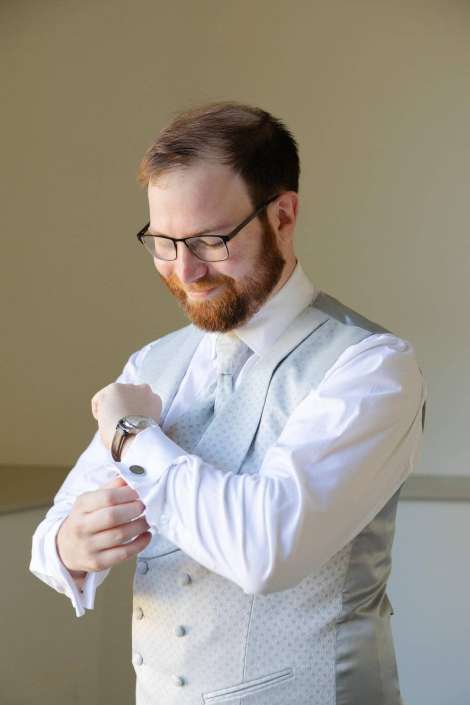 Bearded man wearing glasses, a light gray patterned waistcoat and white shirt, looks down while adjusting his wristwatch.
