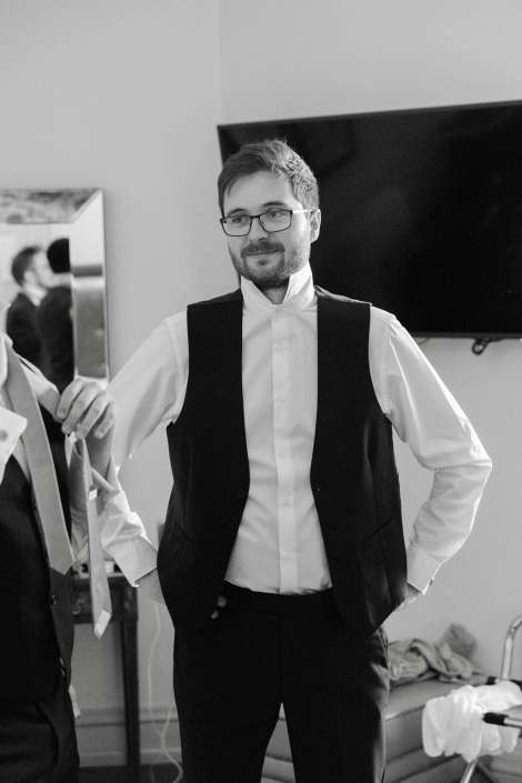 A man in a white dress shirt and black waistcoat stands with hands on his hips, wearing glasses.