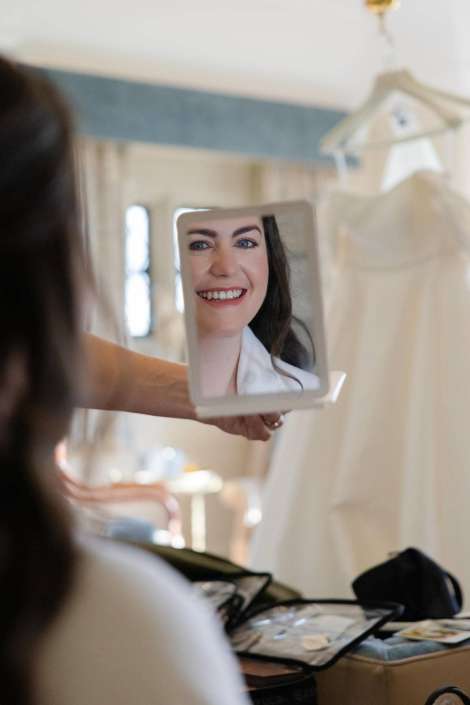 A hand holds a small mirror reflecting a smiling woman, with a wedding dress hanging in the background.