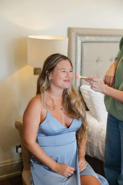 A smiling woman in a light blue dress sits in a bedroom as a makeup artist applies blush with a brush.