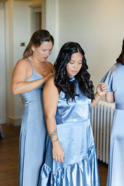 Two women in light-blue satin dresses stand indoors, with one adjusting the other’s shoulder strap.