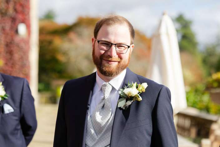 Smiling man with glasses and a beard in a navy suit with a light gray patterned tie and a white boutonniere, standing outdoors.