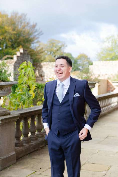 A man in a navy suit with a light blue tie and white pocket square stands on a stone terrace with a balustrade, smiling broadly.