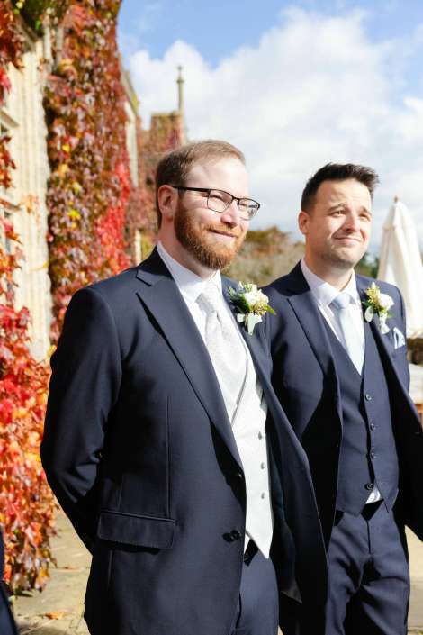 Two men in navy suits with boutonnieres stand outside beside a wall covered in autumn red ivy under a blue sky.