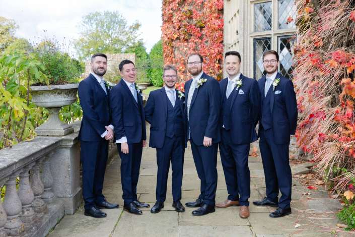 Six groomsmen in navy suits with boutonnieres pose outdoors beside an ivy-covered stone wall in autumn.