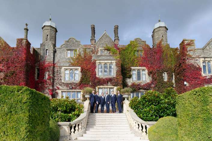 Group of men in dark suits pose on a grand stone staircase in front of an ivy-covered historic mansion.