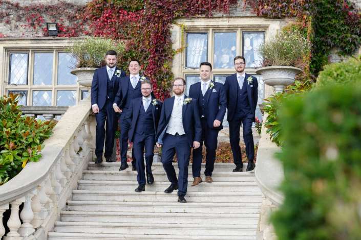 Six men in navy suits pose on a stone staircase outside an ivy-covered historic building with large leaded-glass windows.