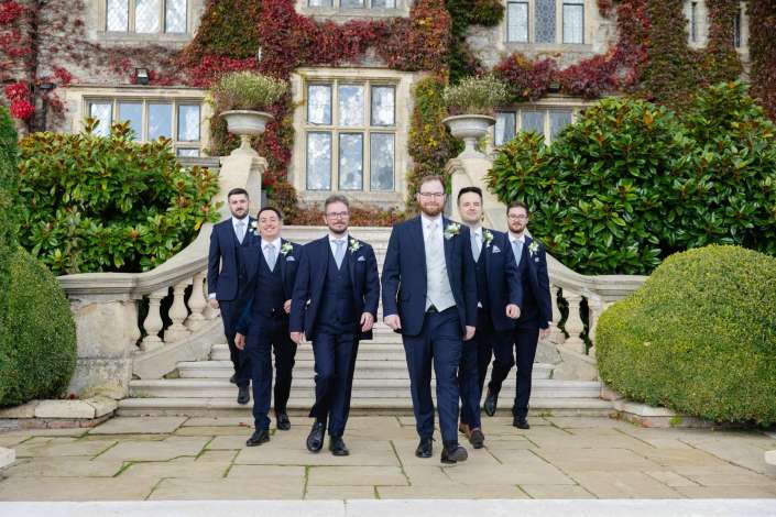 Group of men in navy suits walk down stone steps in front of an ivy-covered historic mansion.