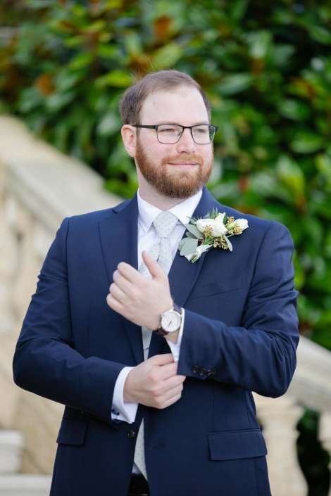 A bearded man in a navy suit with a boutonniere adjusts his cuff outdoors.