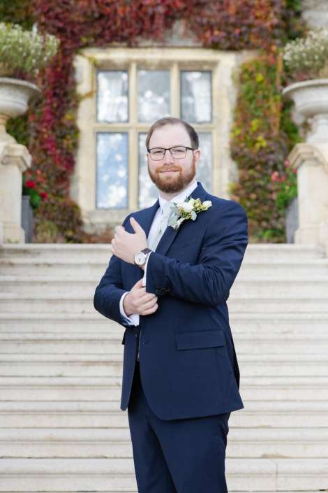 A bearded man in a navy suit with a boutonniere stands on stone steps in front of an ivy-covered building, adjusting his sleeve.